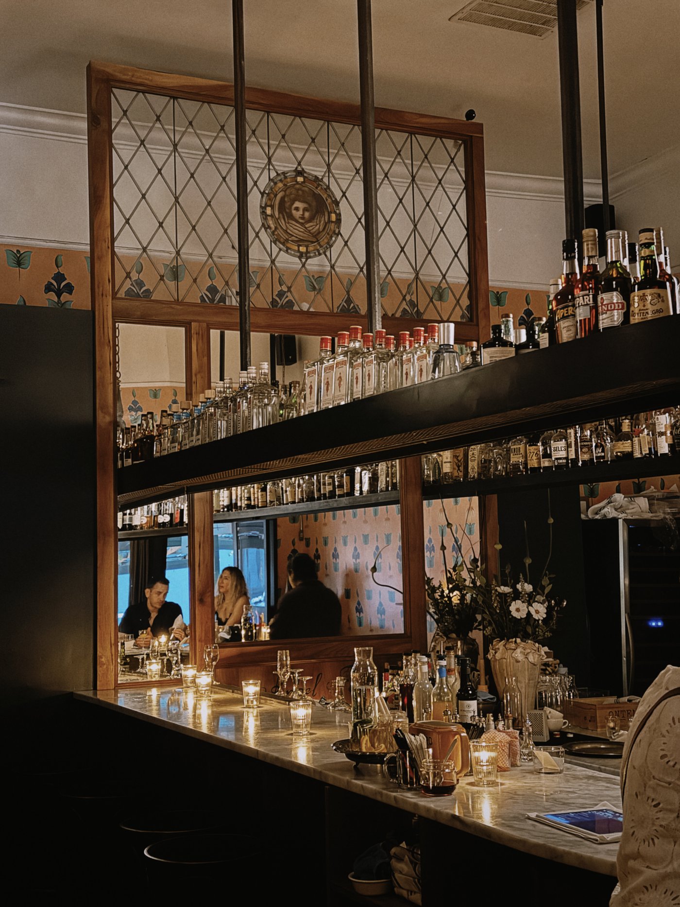An evocative and intimate haunt where flickering candlelight reflects off a white marble bar and tiered spirits. The intricate stained-glass accents and vintage-inspired textures create a soulful, clandestine atmosphere for a late-night rendezvous.