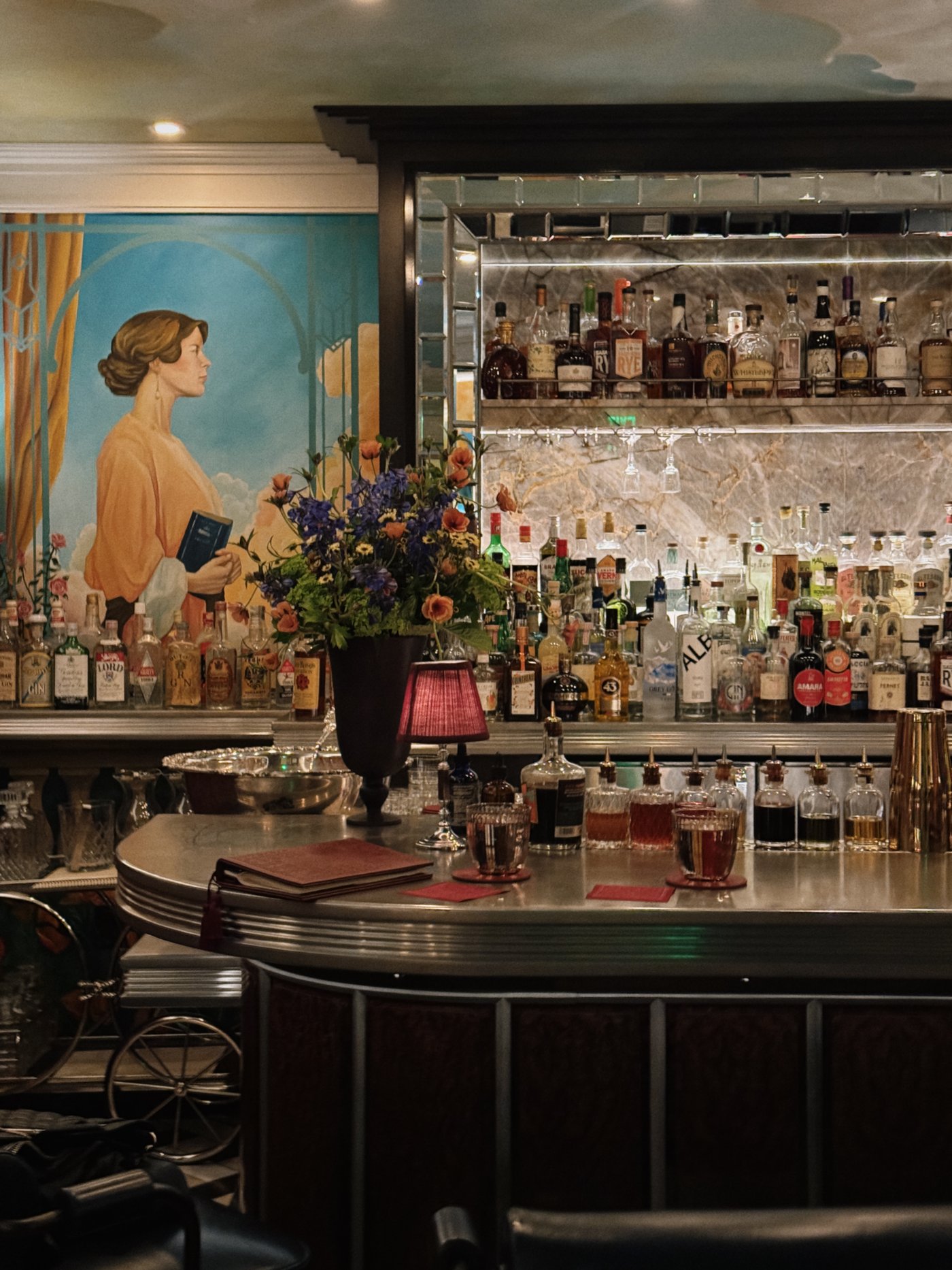 A masterclass in cinematic charm, this intimate bar nook pairs a grand portrait mural with a gleaming pewter top and amber-lit spirits for a transportive drinking experience. The layers of mirrored glass and fresh botanicals lend a storied, romantic soul to the corner.
