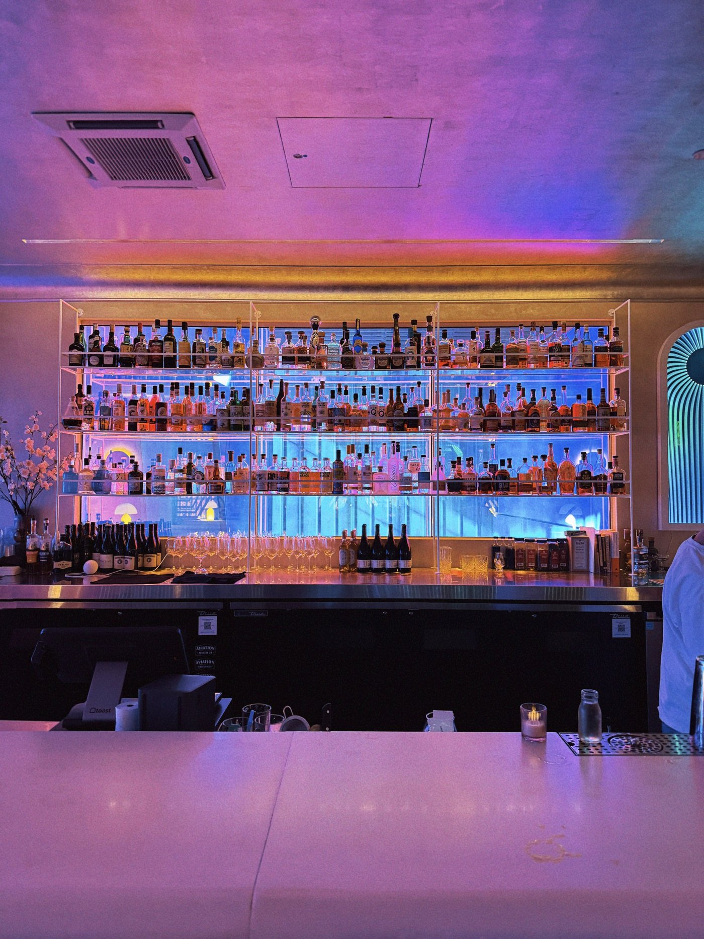 A striking back-lit bar display casts a vaporwave-inspired glow over the room, blending modern minimalism with an electric, high-energy atmosphere. The contrast between the cool blue lighting and the warmth of the spirits creates a magnetic, cinematic backdrop for late-night cocktails.
