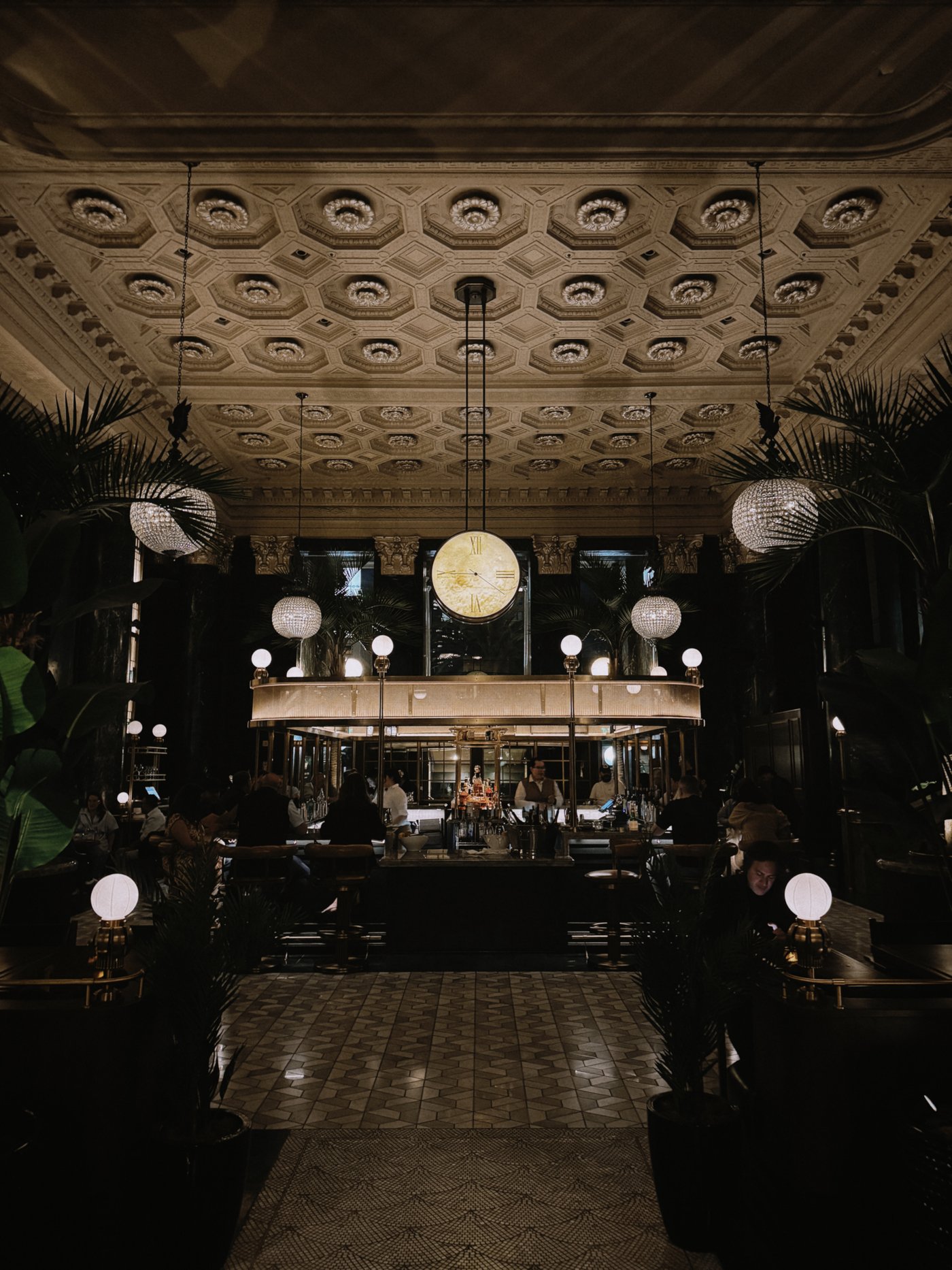 An opulent, subterranean-feeling sanctuary where Gilded Age grandeur meets a lush, tropical-infused cocktail culture. The dramatic coffered ceilings and glowing crystal globes cast a sultry, golden light over the circular brass bar, creating an atmosphere of timeless mystery.