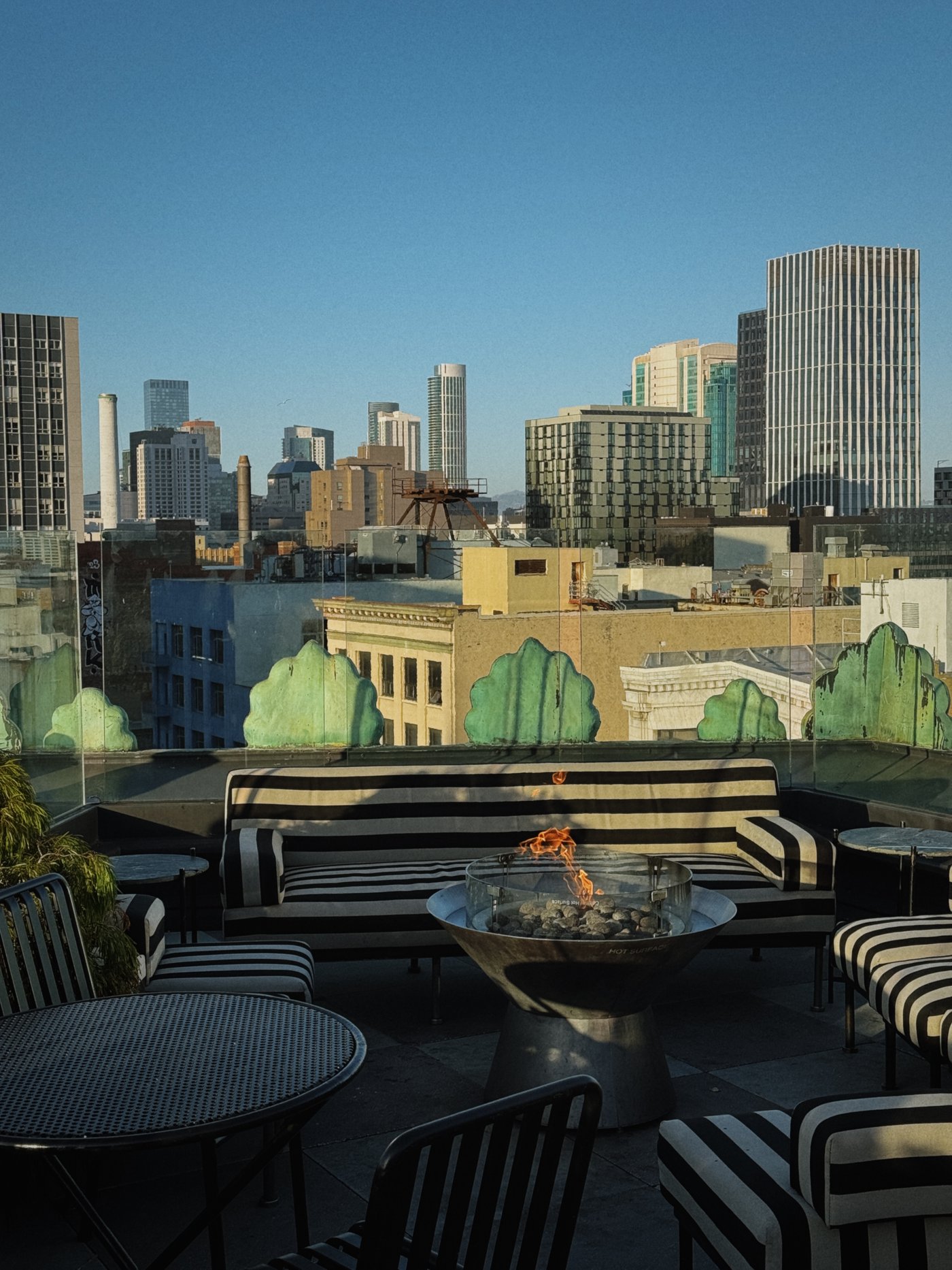 Perched high above the bustling city, this rooftop escape combines bold monochrome stripes with the flickering warmth of an open fire pit. It is an effortlessly chic vantage point where the architecture of the skyline becomes the ultimate backdrop for golden hour cocktails.