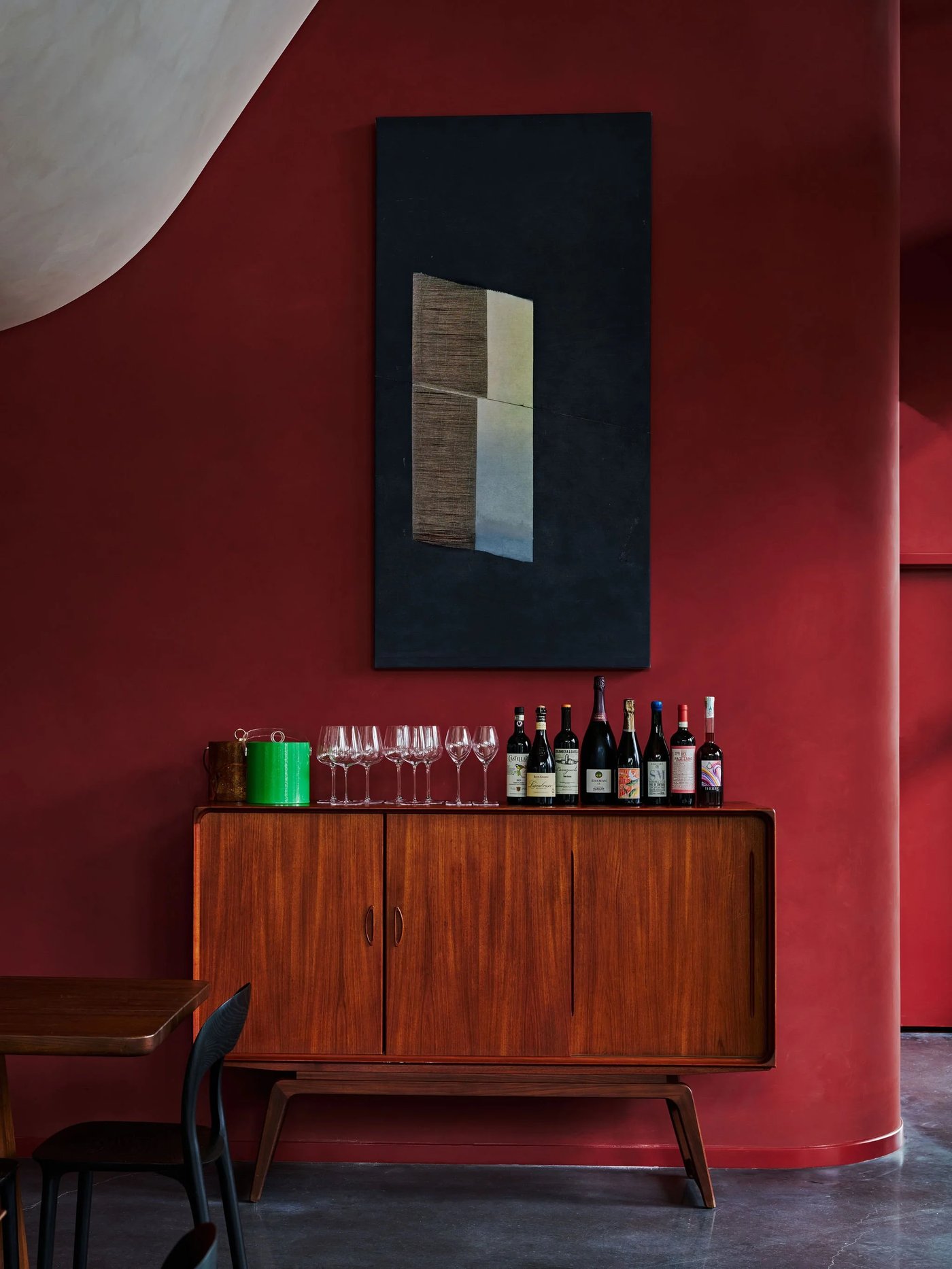 Enveloped in a seductive crimson hue, this corner exudes an air of quiet luxury and intellectual charm, where mid-century design meets a curated selection of fine vintages. The interplay between the textured plaster walls and the minimalist abstract art creates a sophisticated backdrop for an evening of slow sipping and conversation.