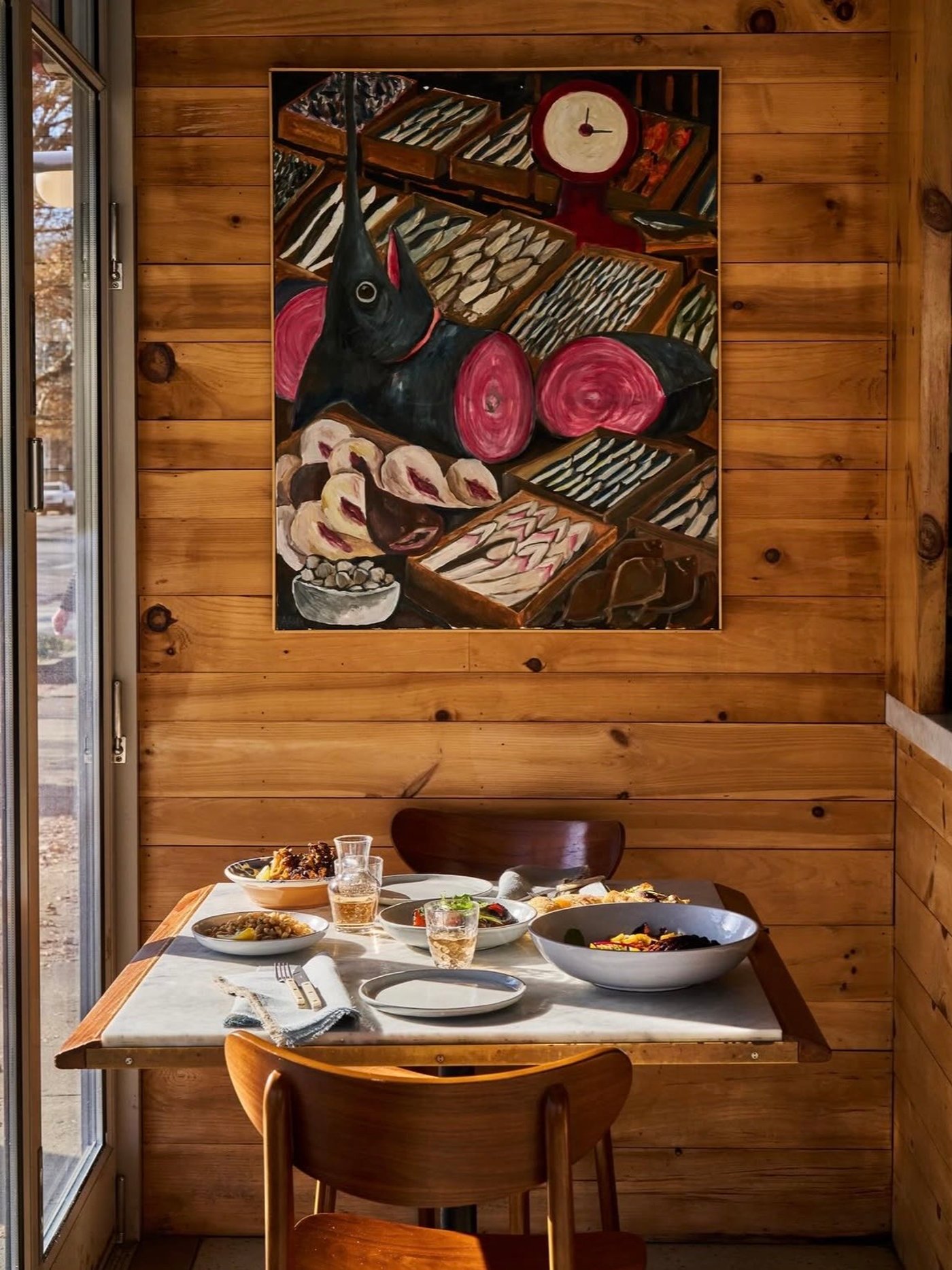 Cloistered in honey-hued pine, this intimate table setting pairs mid-century elegance with a whimsical, fish-market-inspired oil painting for a vibe that is both grounded and artful. Soft daylight washes over marble and ceramic, creating a tactile, sensory environment perfect for a slow, thoughtful lunch.