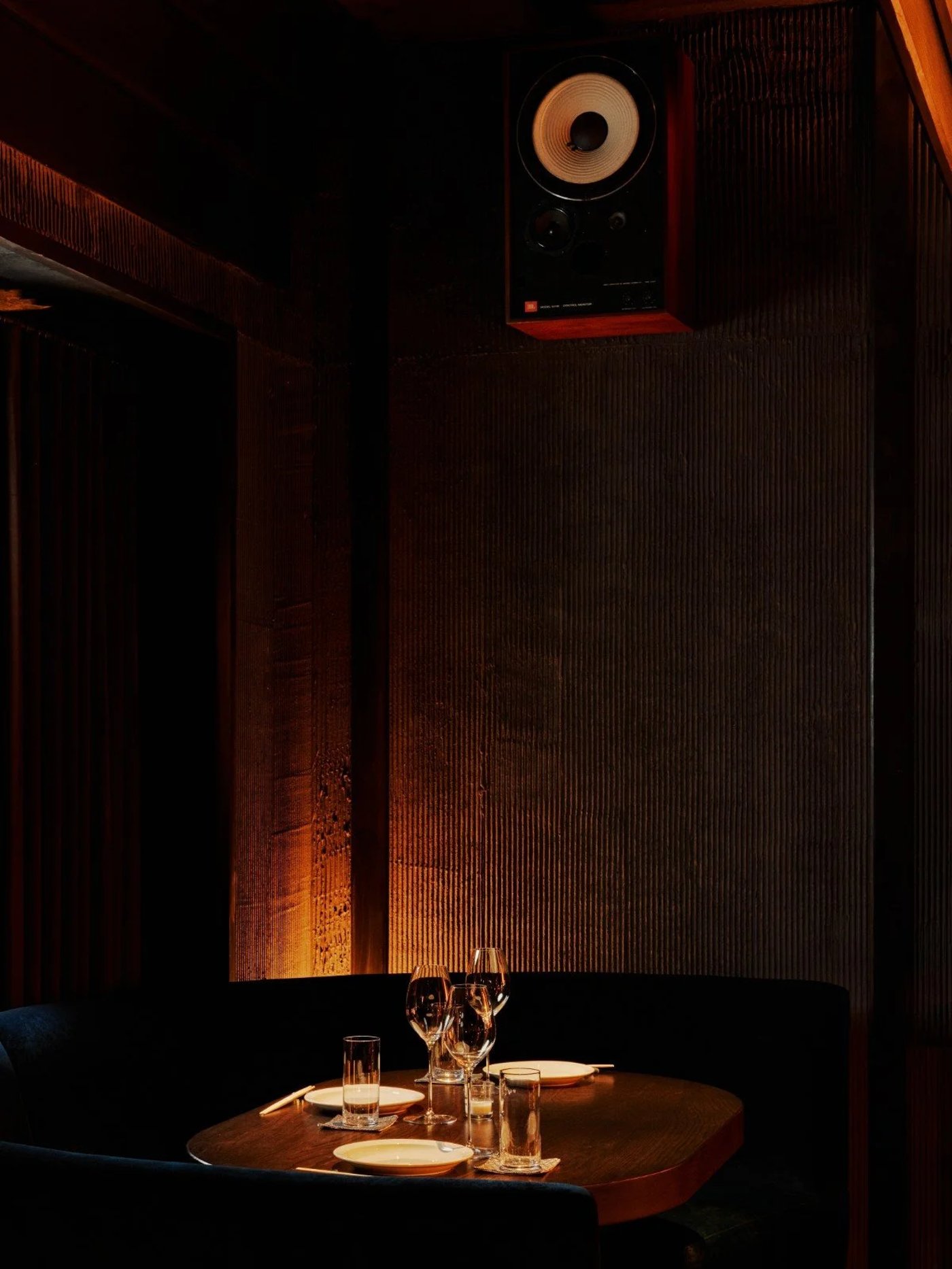 An exercise in shadowed intimacy, this space blends high-fidelity sound with a sultry, low-lit ambiance perfect for late-night secrets. The interplay between the rhythmic reeded walls and the warm pool of light over the table creates a centered, almost cinematic dining experience.