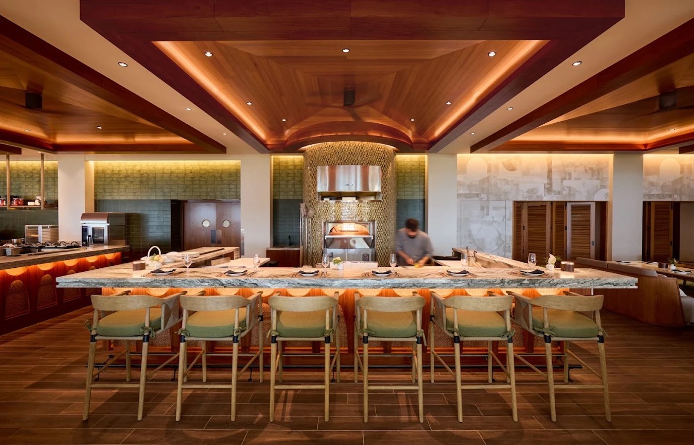 A masterclass in warm architectural symmetry, this open-concept kitchen bar invites guests to witness culinary artistry beneath a stunningly lit coffered wood ceiling. The space balances tactile organic materials like woven cane and sage upholstery with the cool, expansive luxury of a thick marble counter.