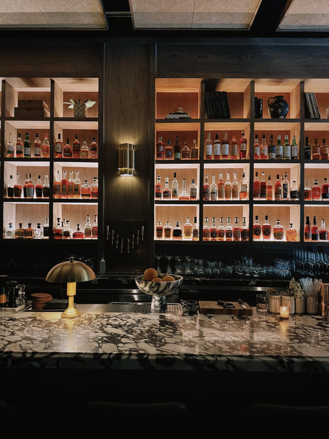 An intimate, amber-hued sanctuary where dramatic marble surfaces meet a meticulously curated library of fine spirits. The warm glow from brass accents and back-lit shelving creates a seductive atmosphere perfect for lingering over a masterfully crafted cocktail.