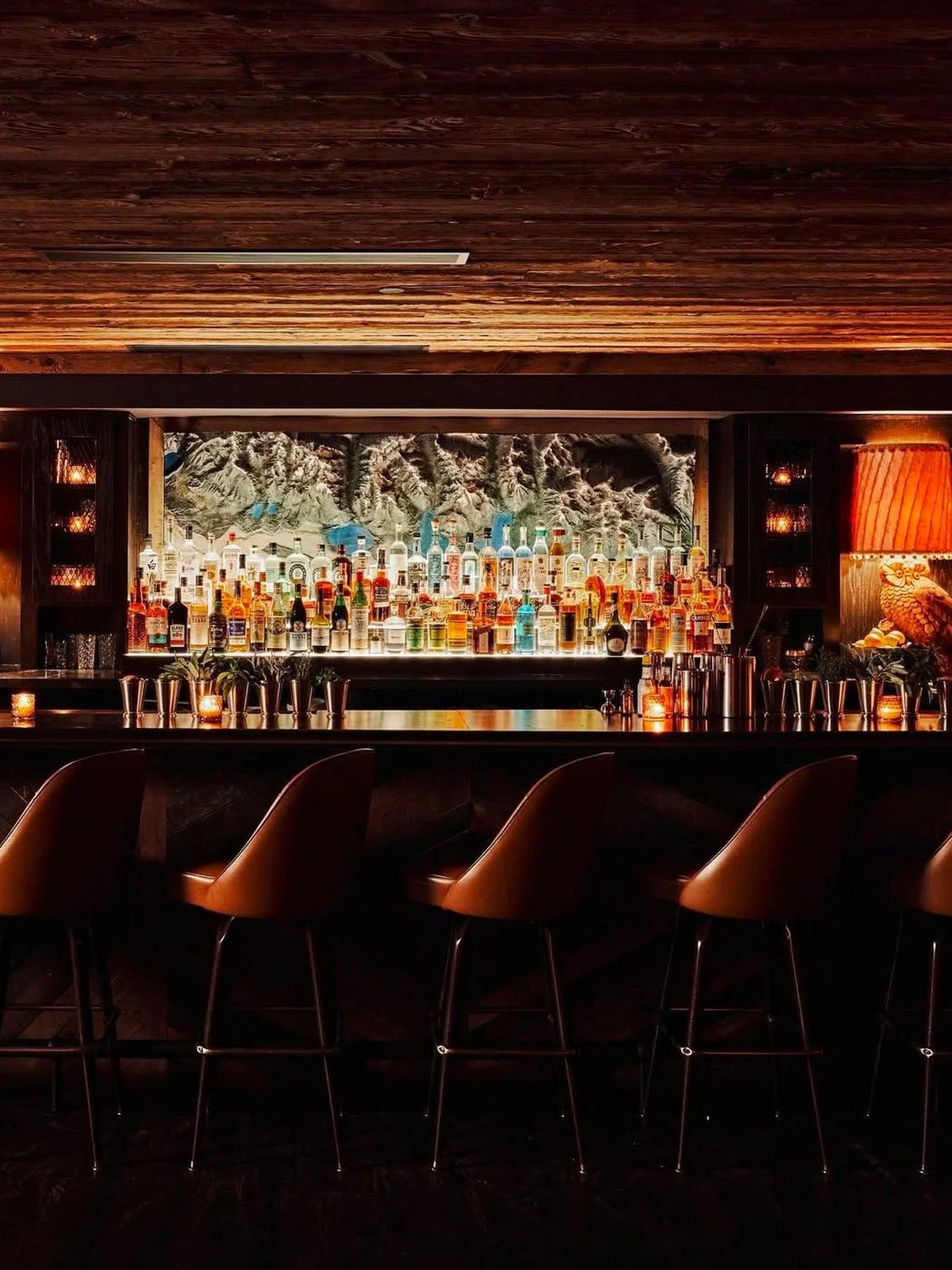 This sultry alpine retreat exudes a moody, après-ski elegance, anchored by a glowingly backlit bar and a stunning topographical mountain mural. The rich interplay of weathered wood textures and butter-soft leather stools creates an intimate sanctuary perfect for long conversations over craft spirits.