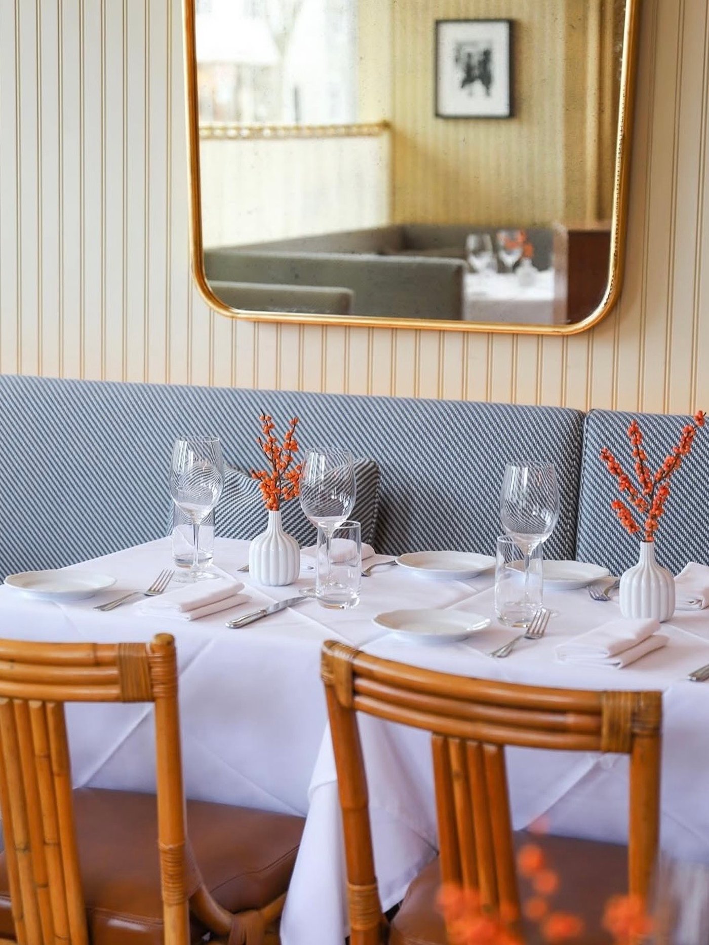 This expertly set corner exudes a timeless charm, where crisp white linens meet the organic warmth of bamboo chairs and subtle coastal stripes. Sunlight bounces off polished crystal and a curved brass mirror, creating a bright, layered setting perfect for a slow, sophisticated lunch.
