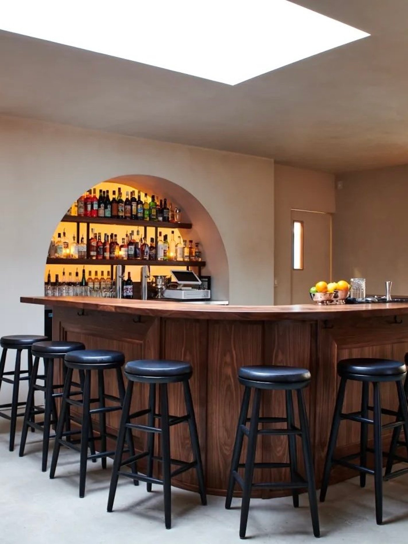 A masterclass in soft minimalism, this cocktail bar pairs the organic warmth of curved walnut with lime-plastered walls and a glowing arched back-bar. The space feels like a serene, hidden hideaway where light from a large skylight illuminates the precision of the architecture.