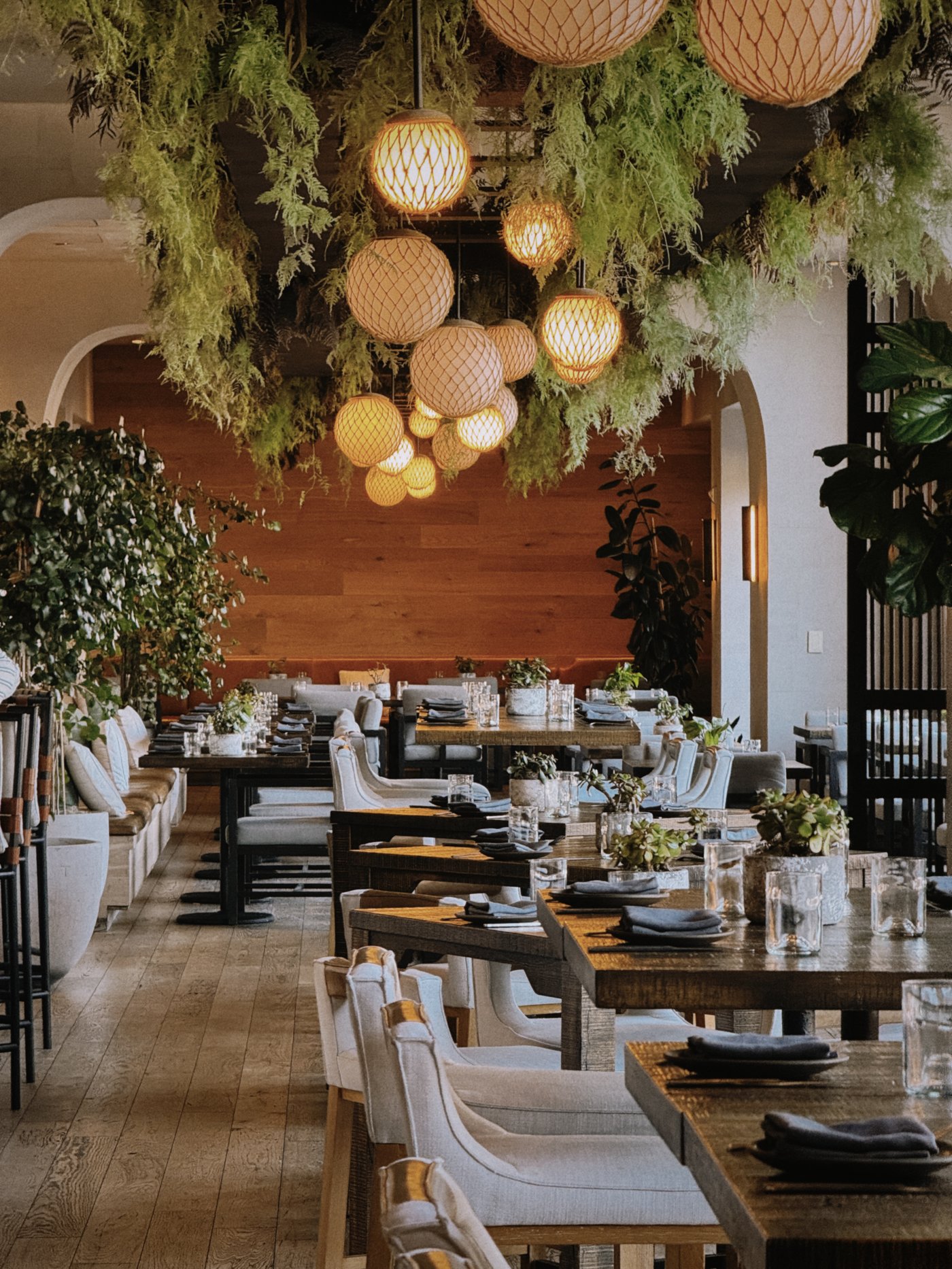 An ethereal biophilic escape where warm, orb-like lanterns nestle within lush greenery to create a glowing canopy above sophisticated wooden tables. The space feels like a secret garden reimagined through a lens of refined, modern luxury.