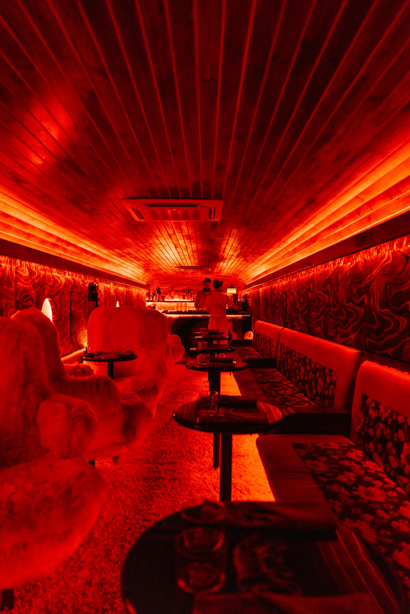 Drenched in a bold crimson glow, this subterranean-style lounge blends vintage tactile patterns with futuristic neon for a surreal, cinematic drinking experience. The mix of shaggy textures and wooden slat architecture creates an intimate, high-fashion sanctuary that feels entirely disconnected from the outside world.