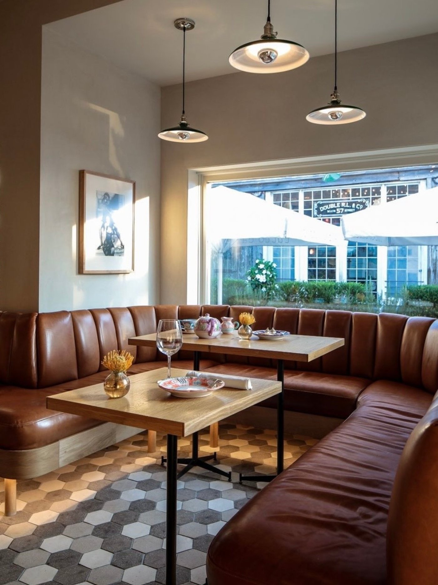 Natural light pours over an expansive cognac leather banquette, creating a warm, textural sanctuary for lingering conversation. The interplay of geometric floor tiles and minimalist wooden tables establishes a perfectly balanced, modern brasserie aesthetic.